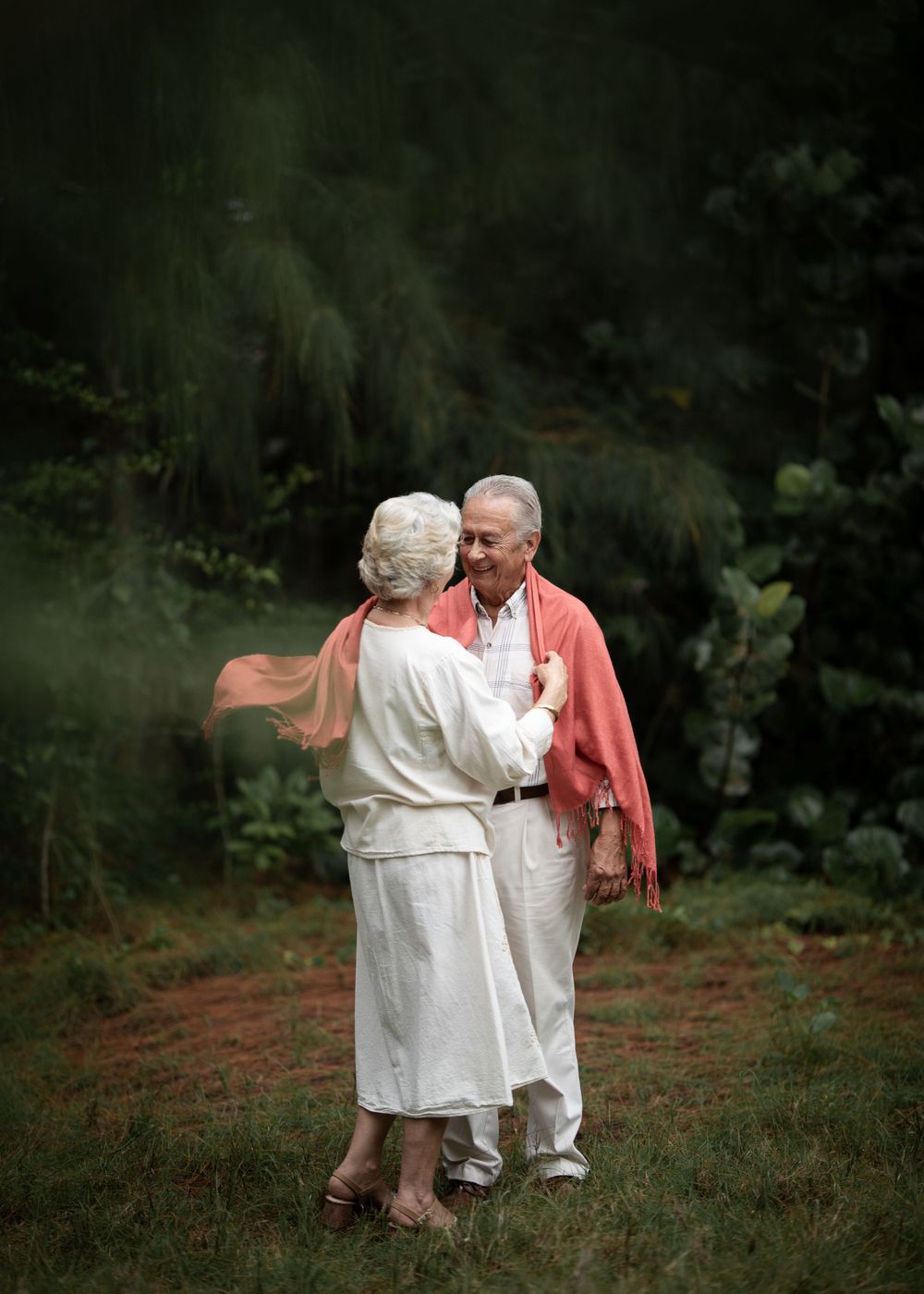 A senior couple embraces while dancing outdoors, with one wearing a coral sweater.