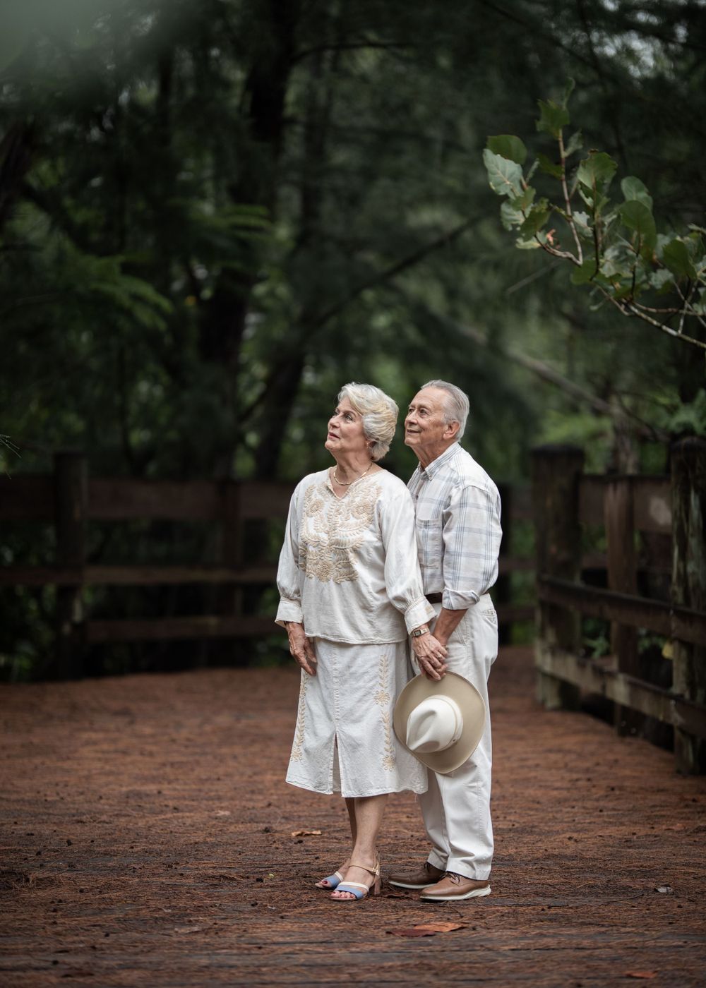 A couple in light colored clothing shares a moment on a wooden bridge in a forest setting.