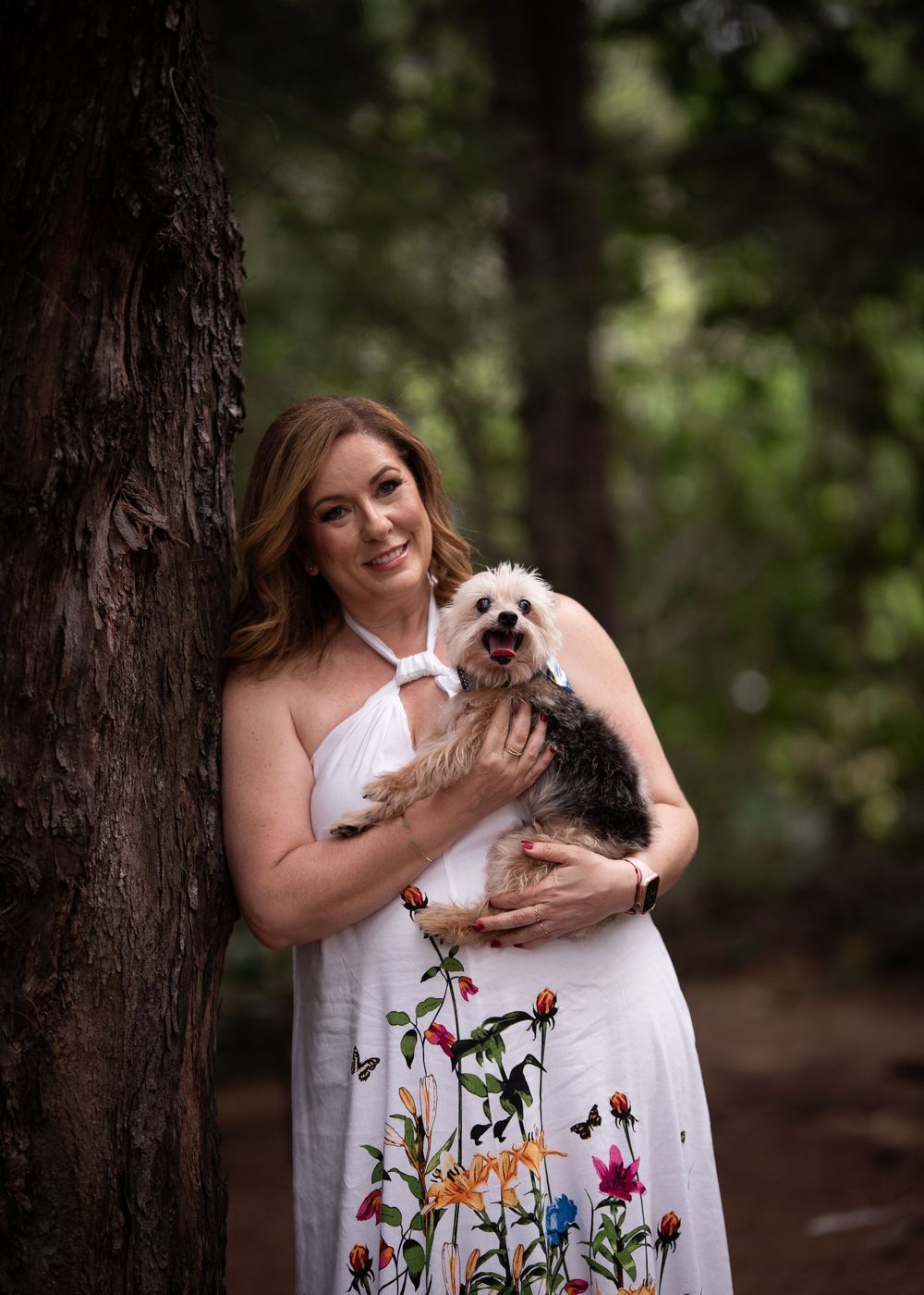 A woman in a floral dress holds a small terrier dog while standing next to a tree.