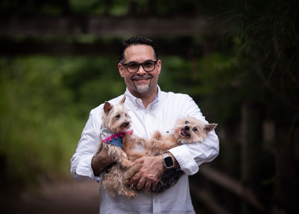 A person in glasses holds two small dogs while standing on a wooded path.