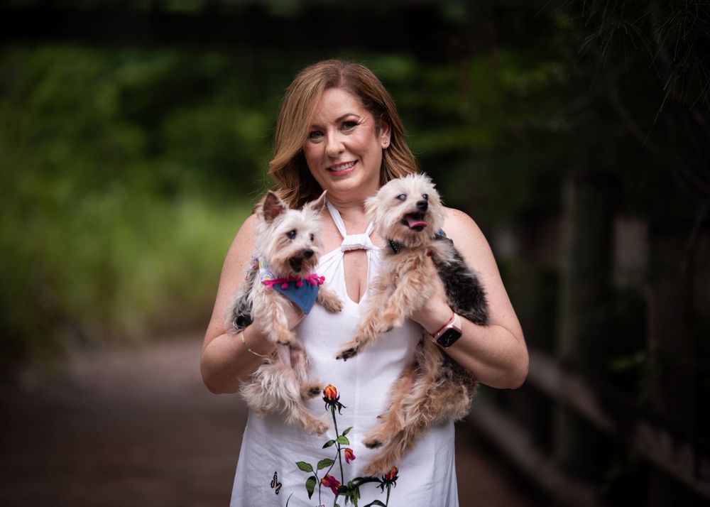 A white floral dress-wearing person holds two small dogs on a wooded path.