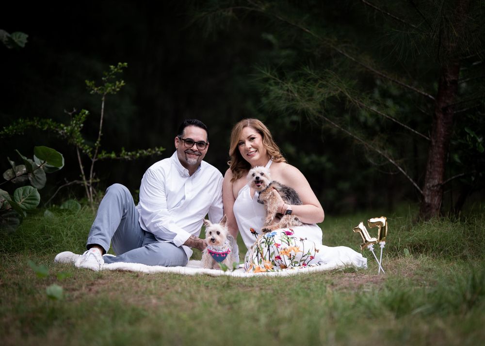 A couple sits on a picnic blanket in the woods with their small dog.