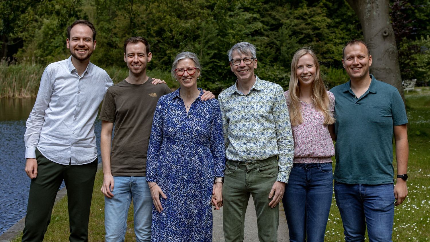 Six people lined up for an outdoor group photo with trees and water behind them.
