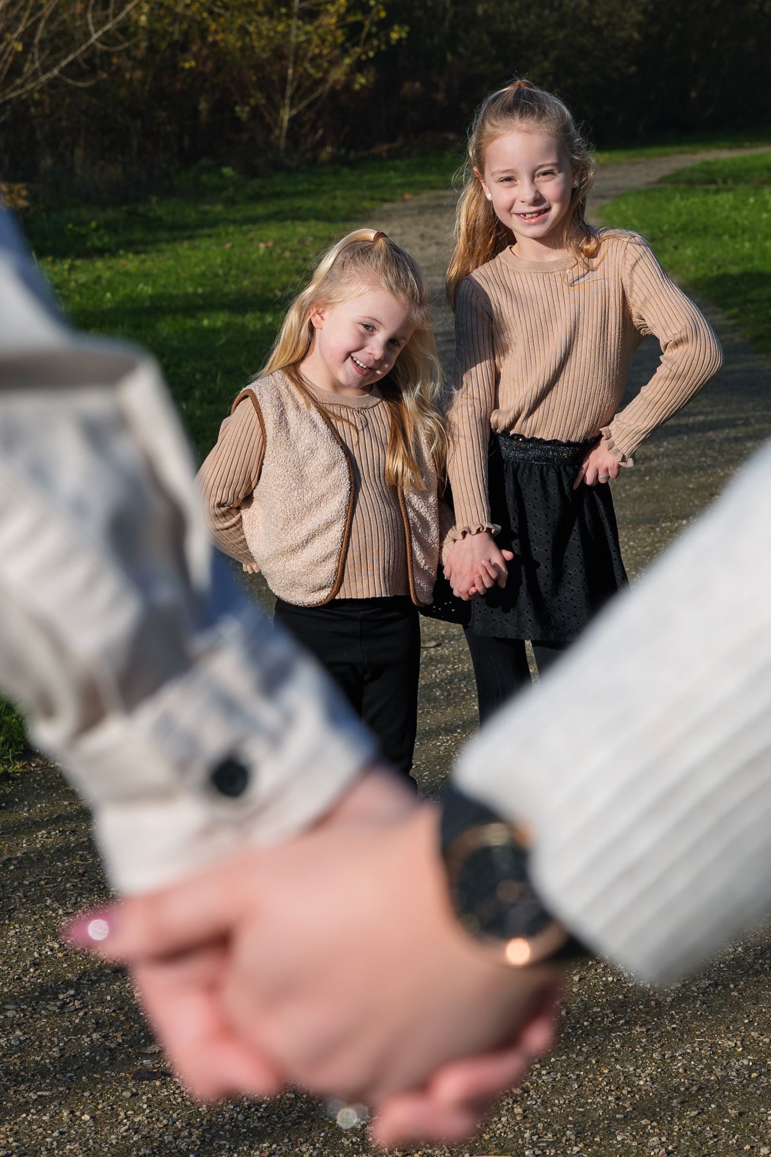 Young children in matching beige sweaters smile together outdoors while hands hold a camera in the foreground.