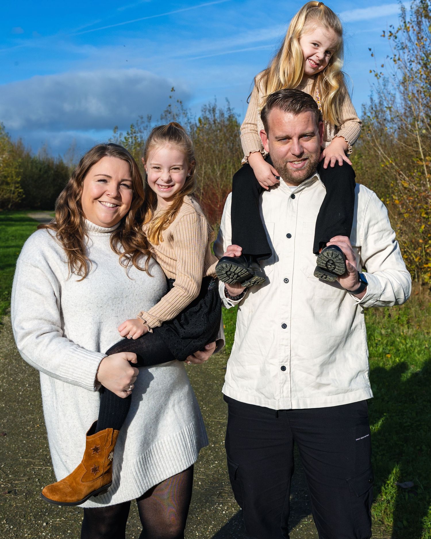 A family poses outdoors on a sunny day with trees and a blue sky in the background.