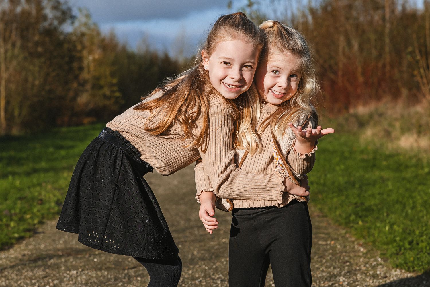 Two young friends wearing matching beige sweaters laugh together on a grassy path during autumn.