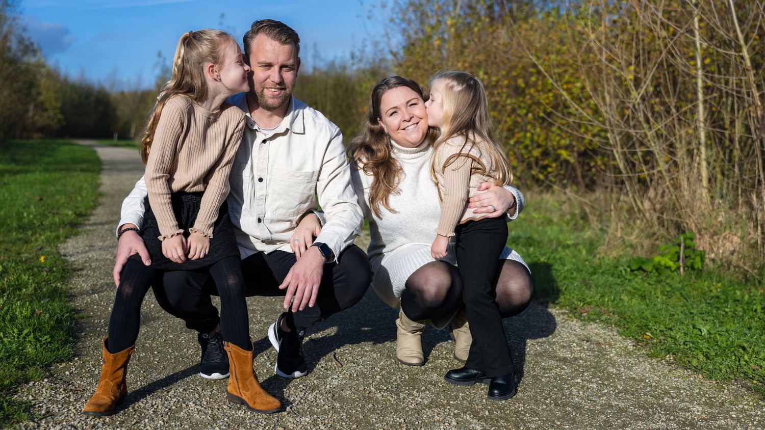 Family of four poses together outdoors on a sunny day along a nature path with spring foliage in the background.