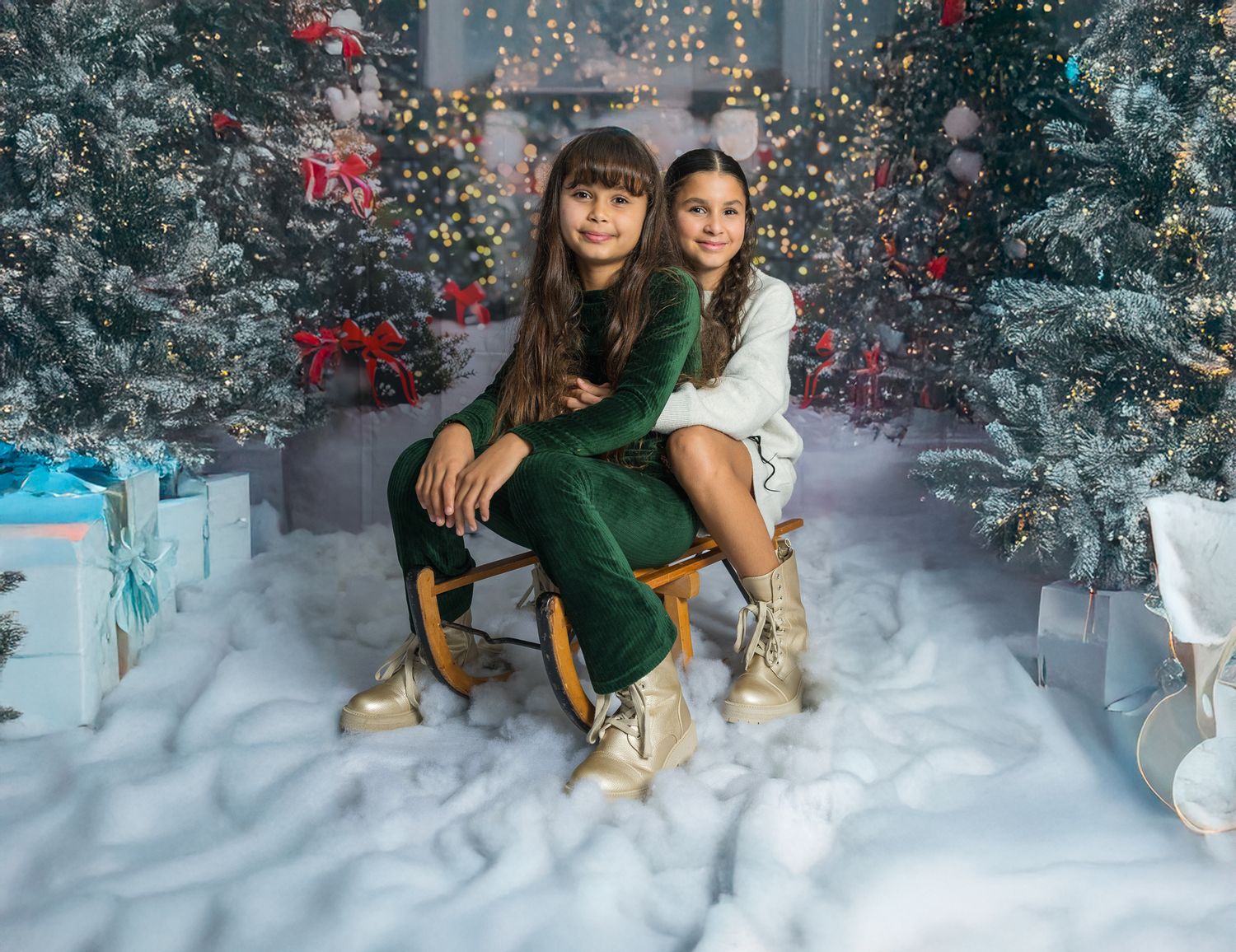 Two people pose on a chair in a winter wonderland setting with snow, Christmas trees and twinkling lights.