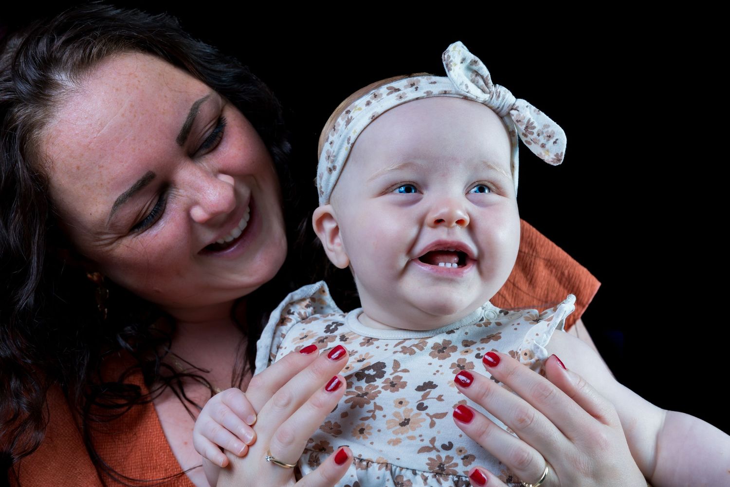 A joyful baby wearing a floral dress and bow headband smiles while being held against a dark background.

- BU Photography -