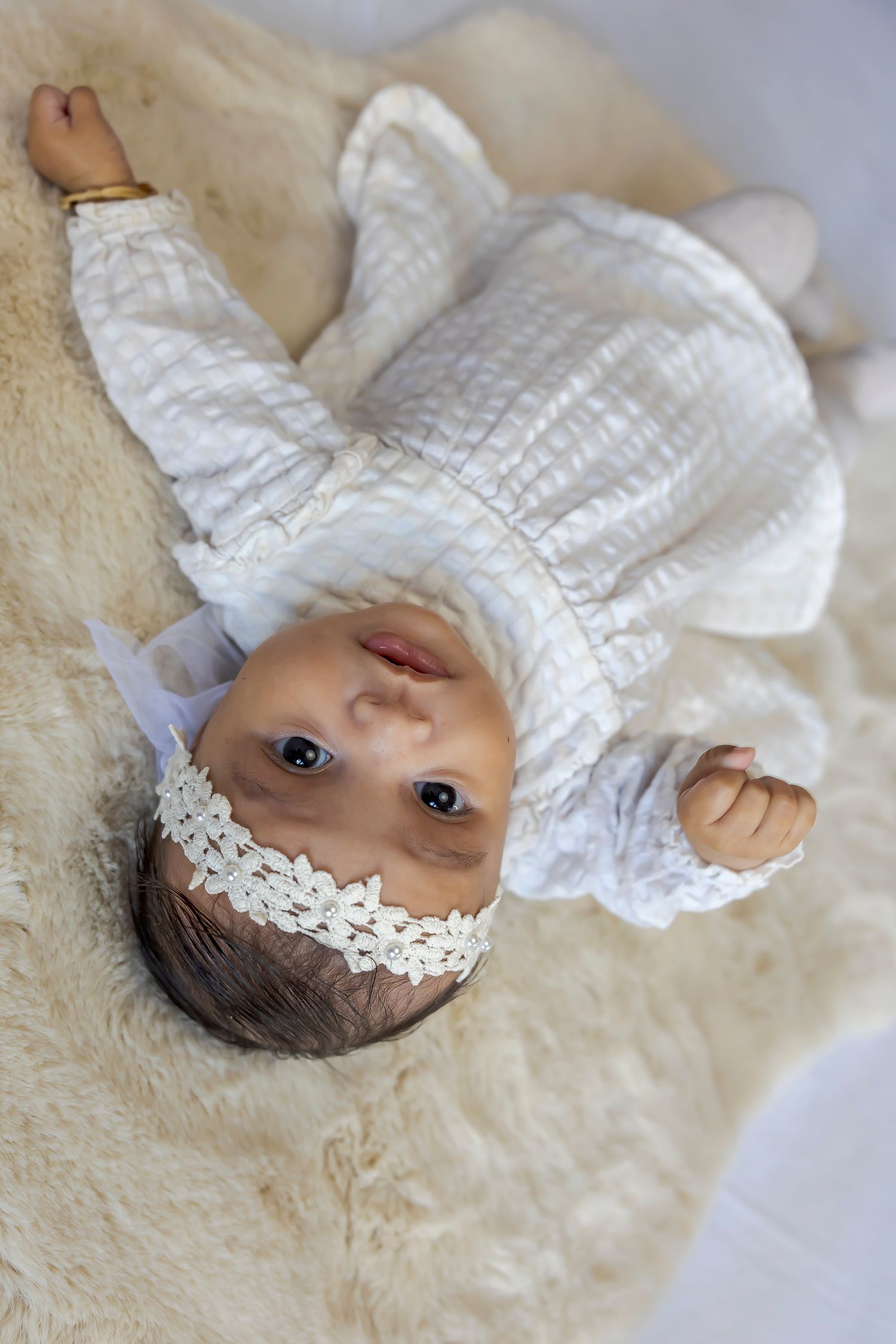 A baby wearing a white dress and floral headband lying on a cream colored blanket.