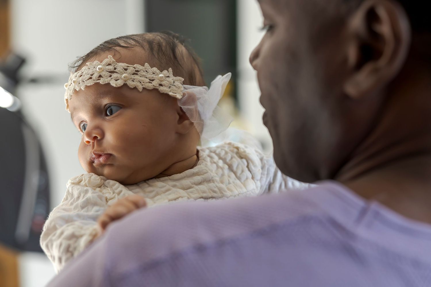 A small baby wearing white lace and a headband being held.