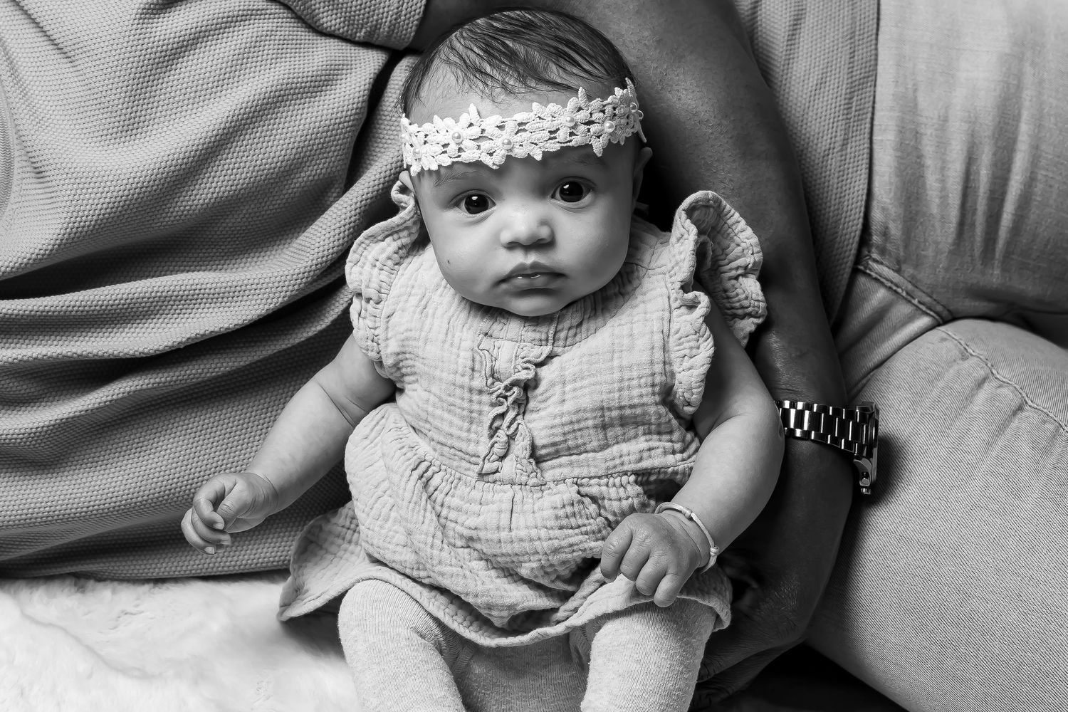 Baby wearing a lace headband and ruffled dress sits on a couch in black and white portrait.