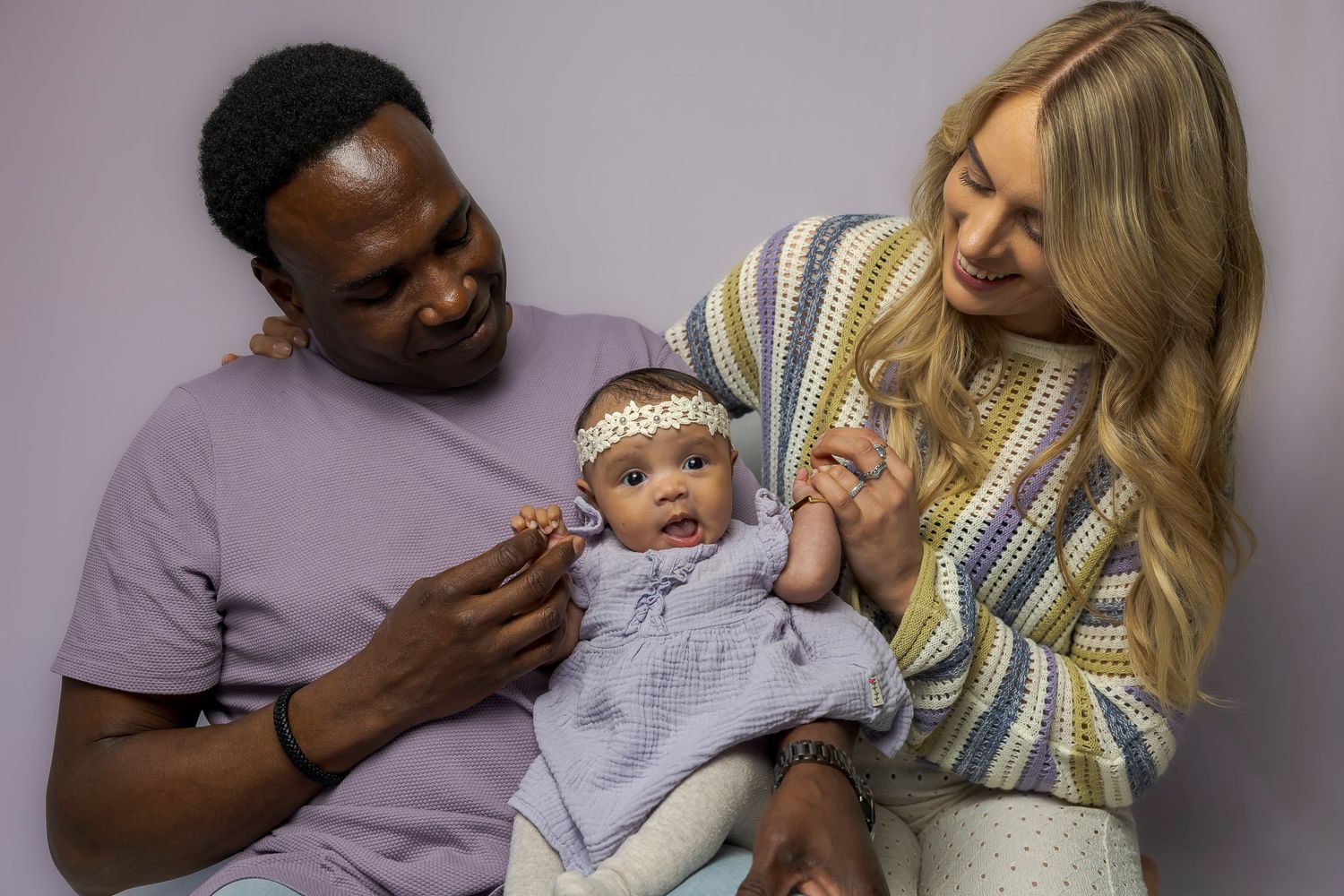 Parents share joyful moment with baby wearing lace headband and blue outfit.