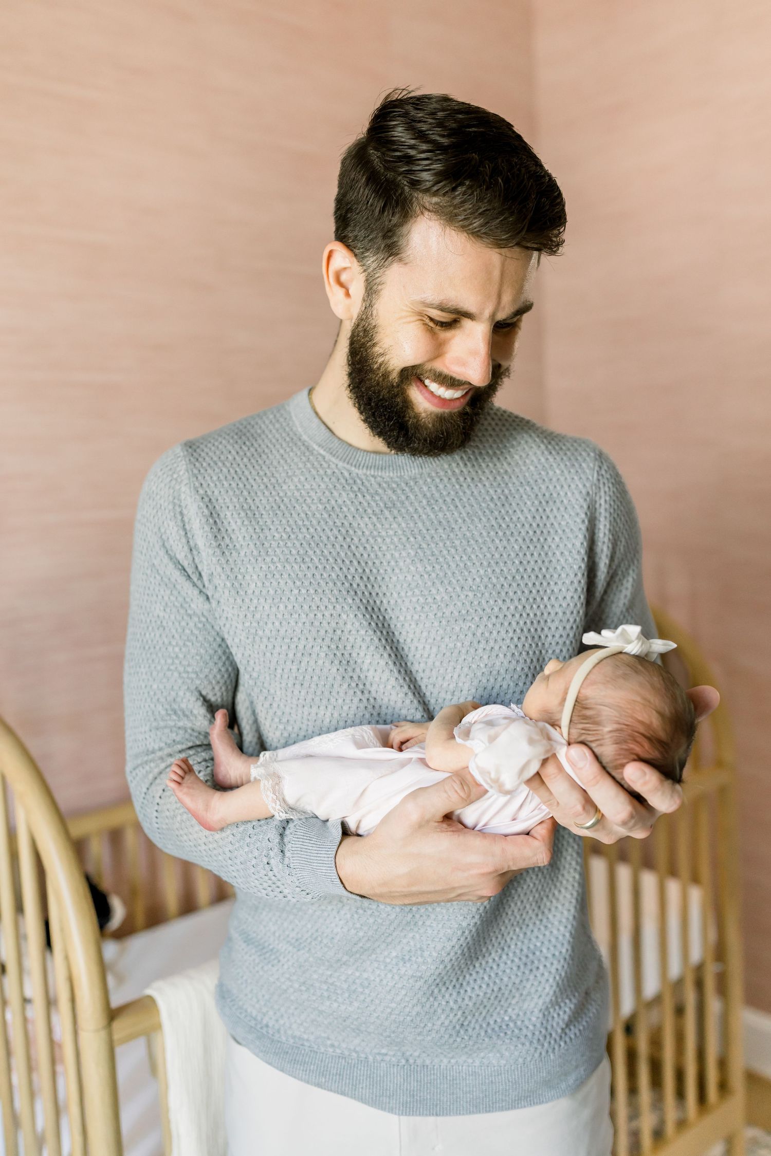 A bearded man in a gray sweater lovingly holds and gazes at a newborn baby in a nursery setting.
