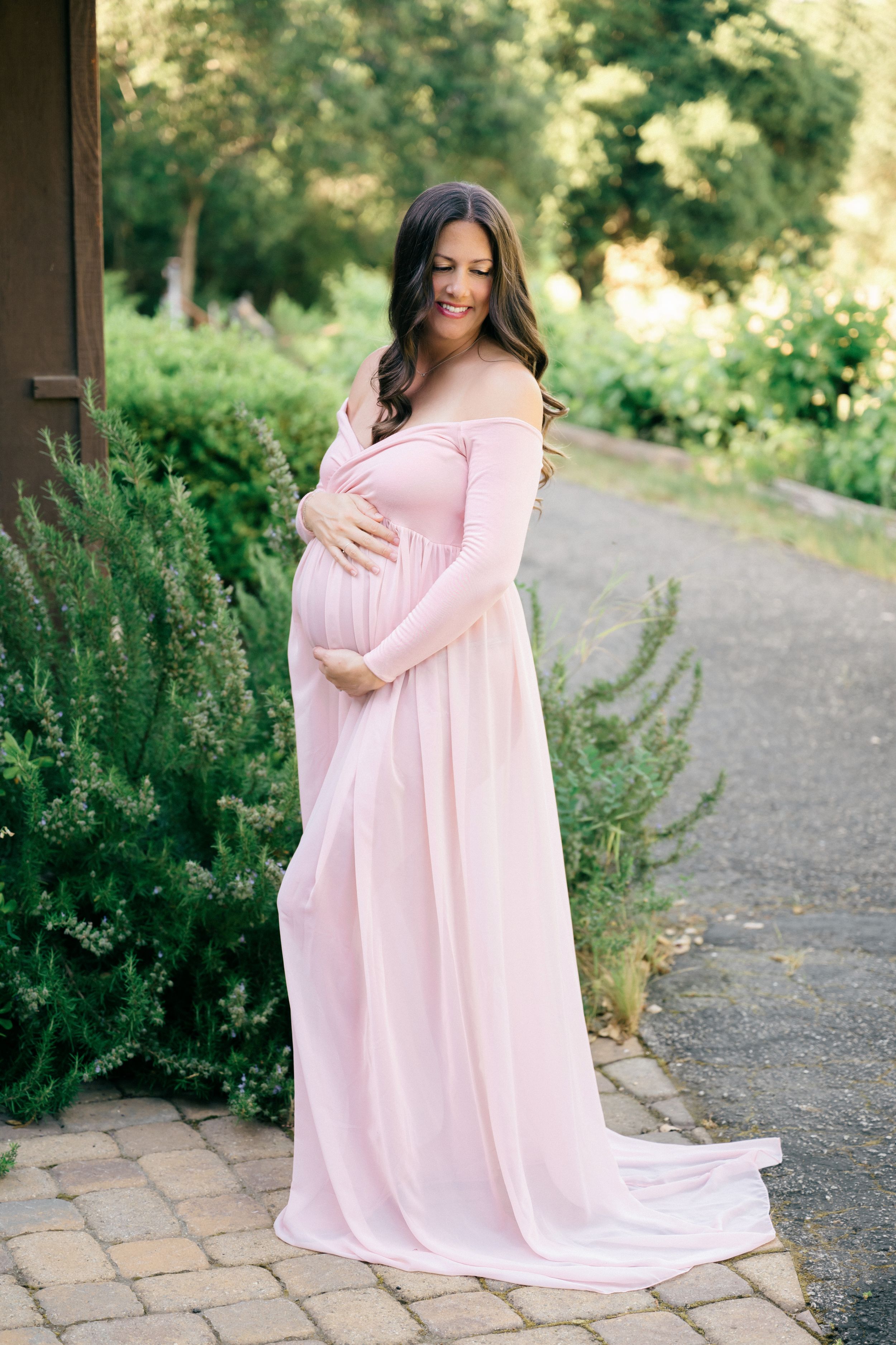 Pregnant woman in elegant pink off-shoulder maternity gown posing outdoors among lush greenery on a brick pathway.