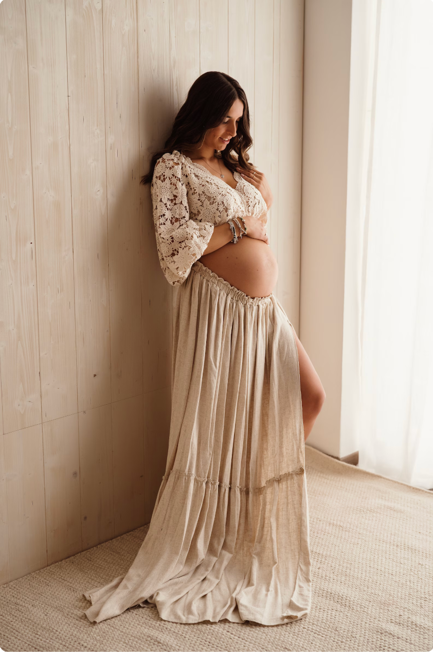 Pregnant woman in elegant cream colored lace dress stands by window in natural light creating ethereal maternity portraits.