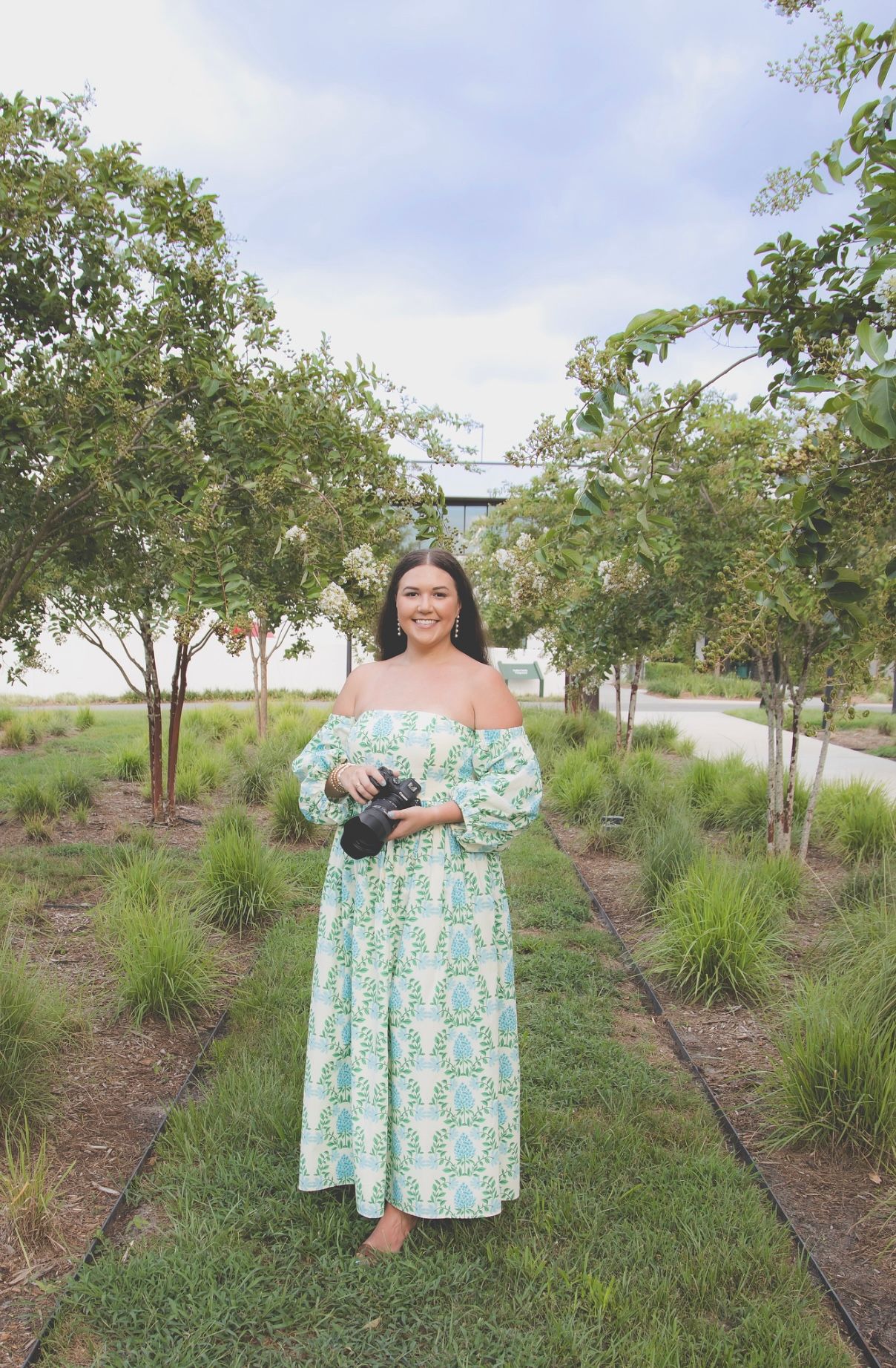 Person in floral off-shoulder dress poses with camera in landscaped garden setting.