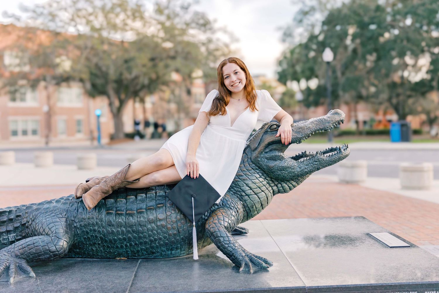 Woman in white dress poses playfully on alligator statue in brick courtyard.