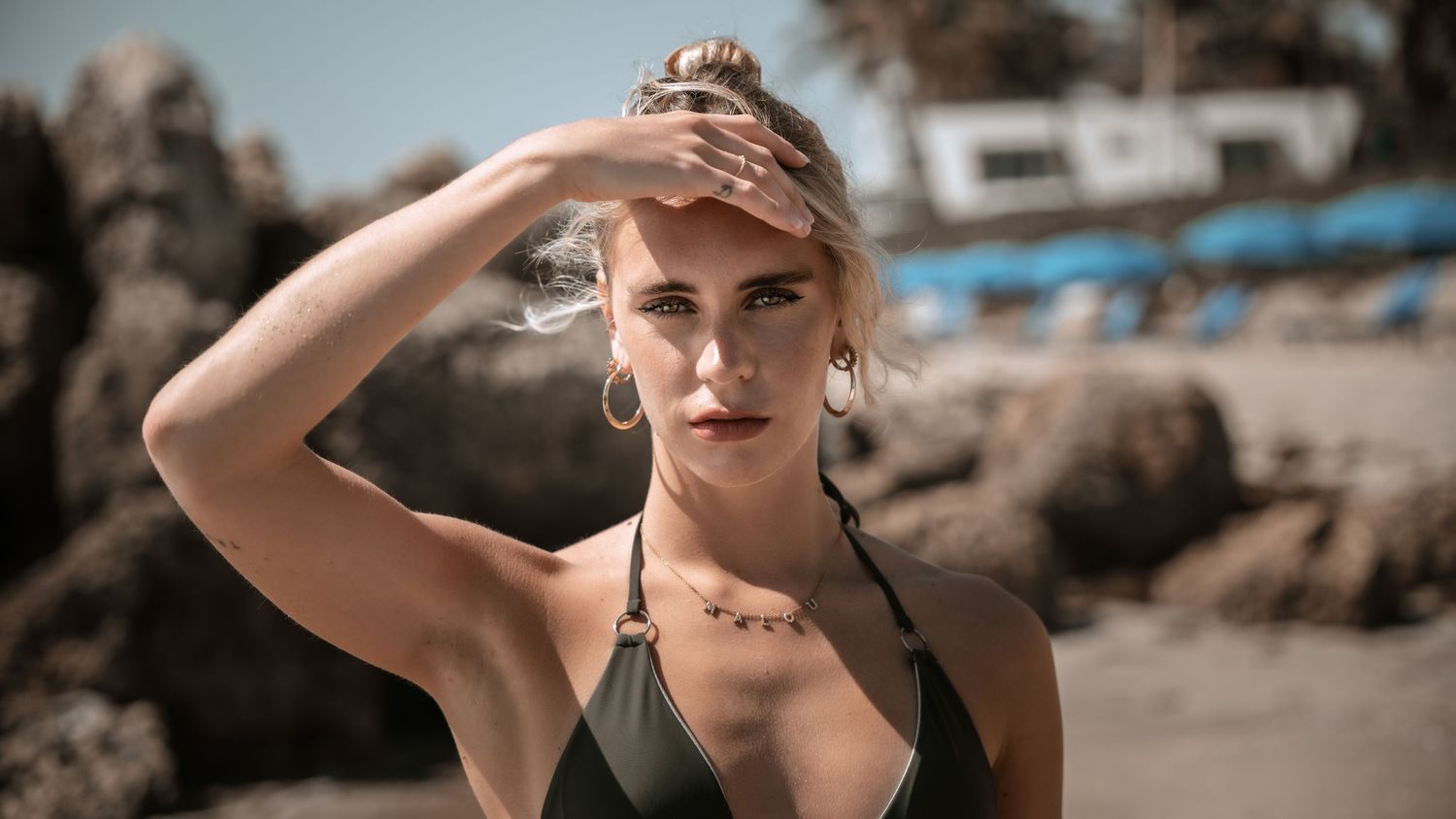 Summer beach portrait with hand shielding eyes against rocky coastal background.