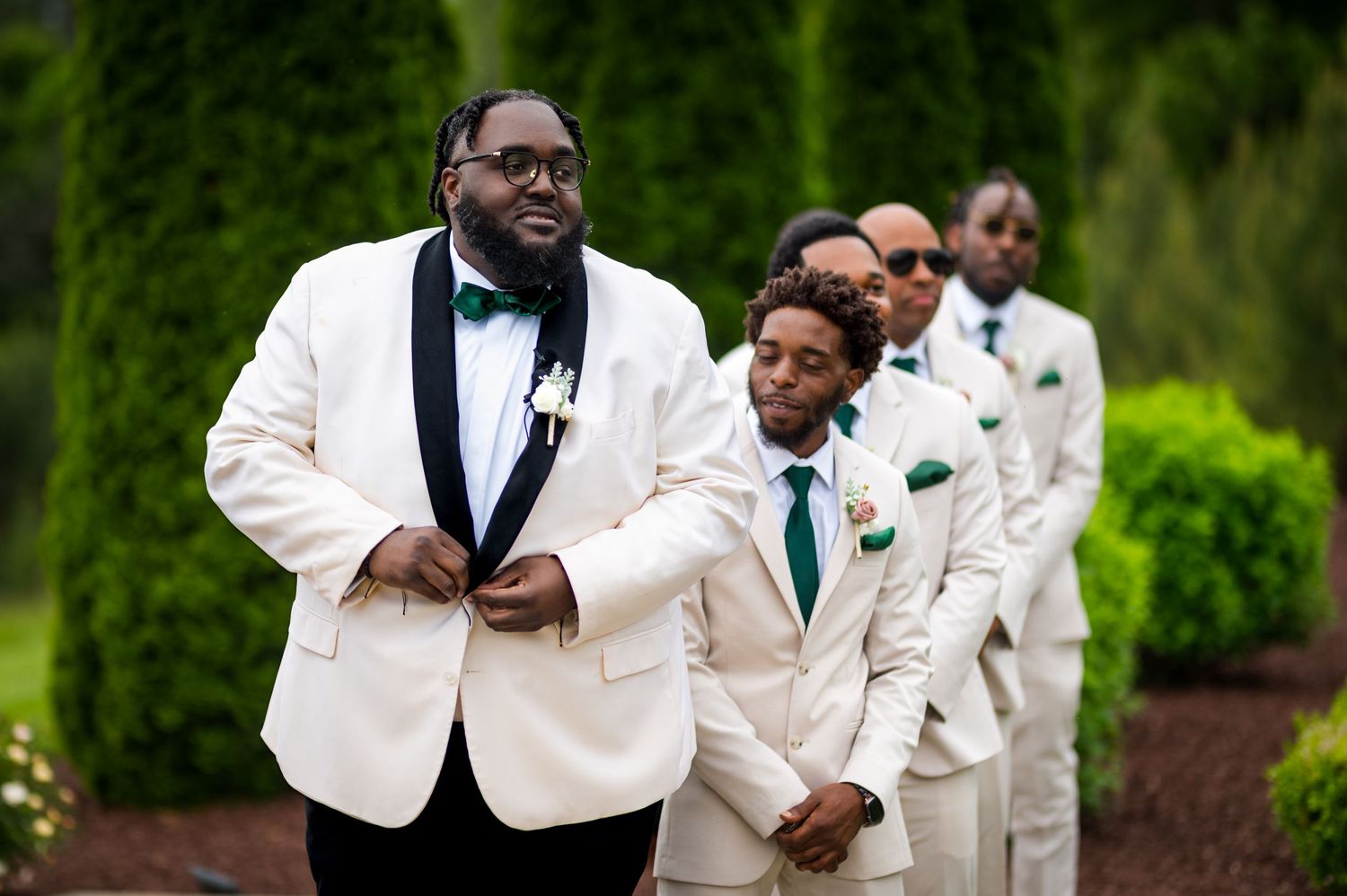 Wedding party in white tuxedos and emerald accents stands together during outdoor ceremony.