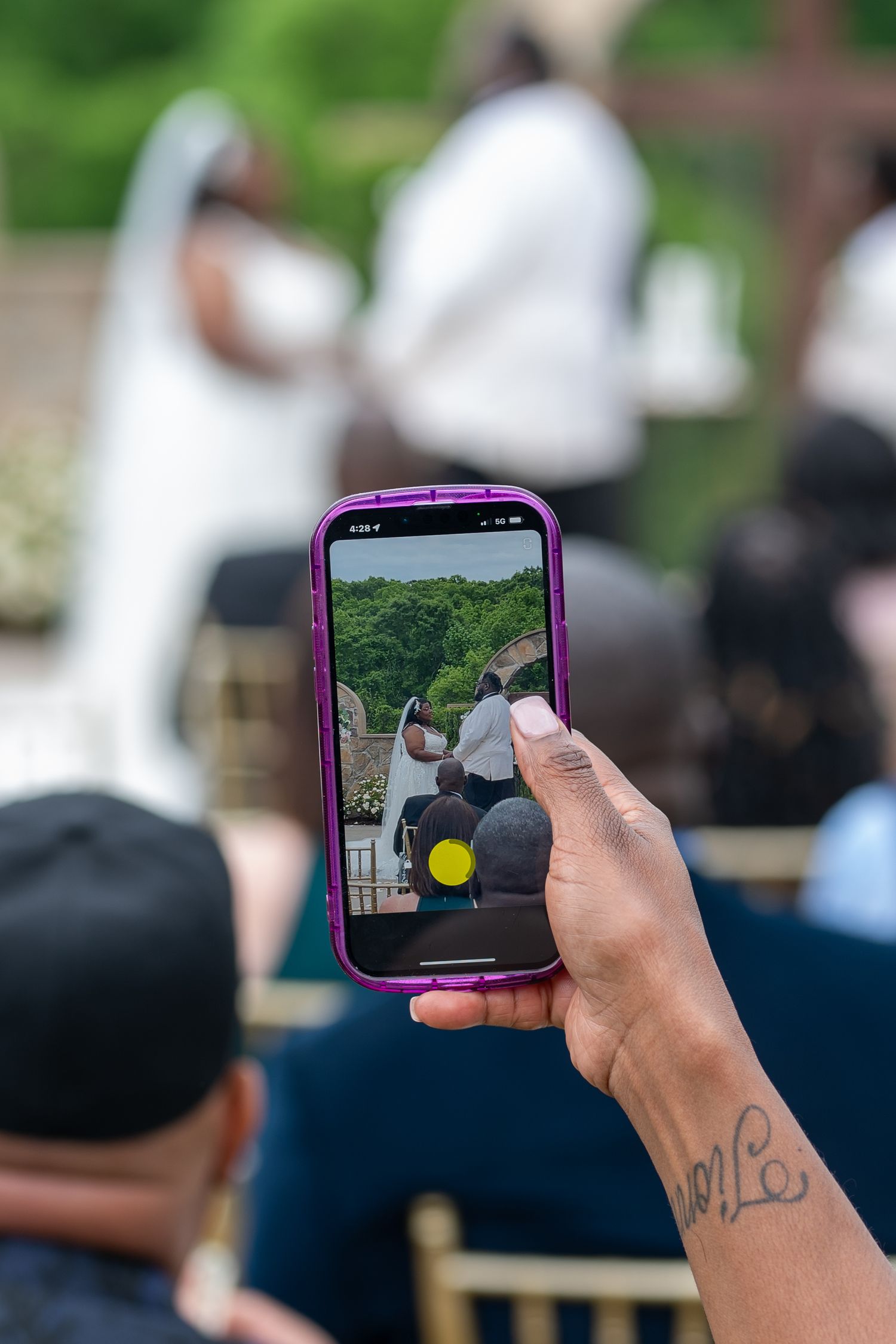 Guest captures wedding ceremony on smartphone with purple case during outdoor celebration.