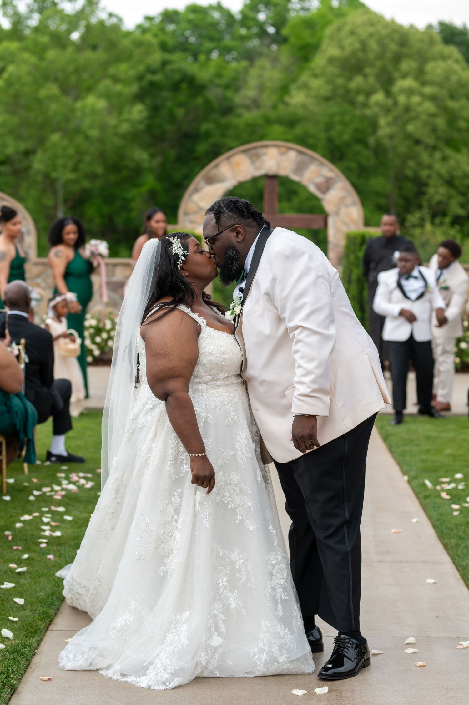 Romantic wedding kiss under garden arch with flower petals scattered on pathway.