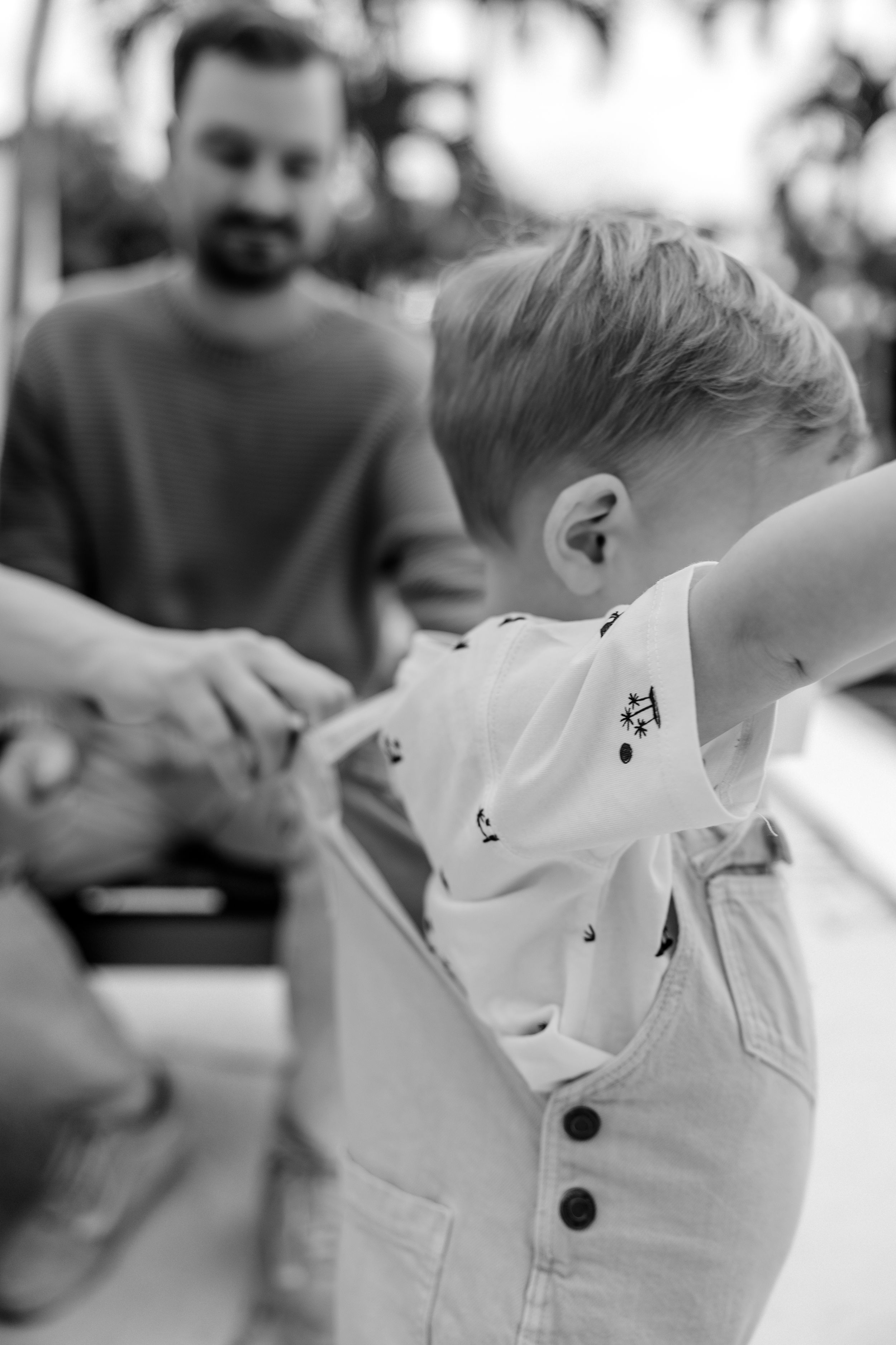 Black and white candid photo of a toddler in a white outfit exploring while being supervised.