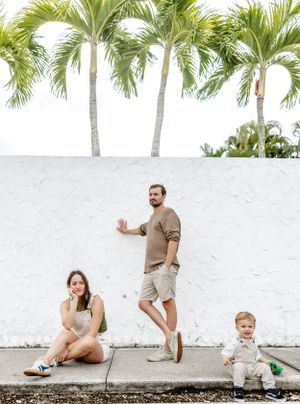 Family poses casually against white wall with tropical palm trees, wearing neutral colored clothing.