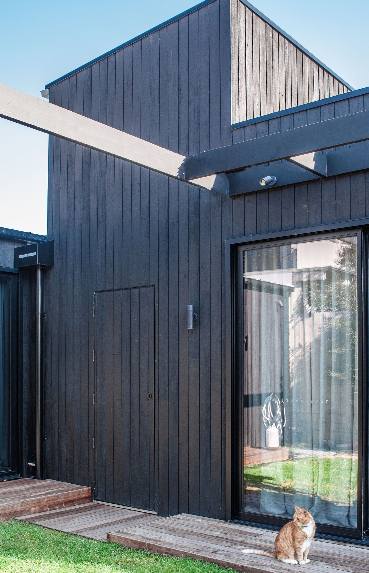 Modern black-clad house exterior with sliding glass door and wooden deck where a ginger cat sits.