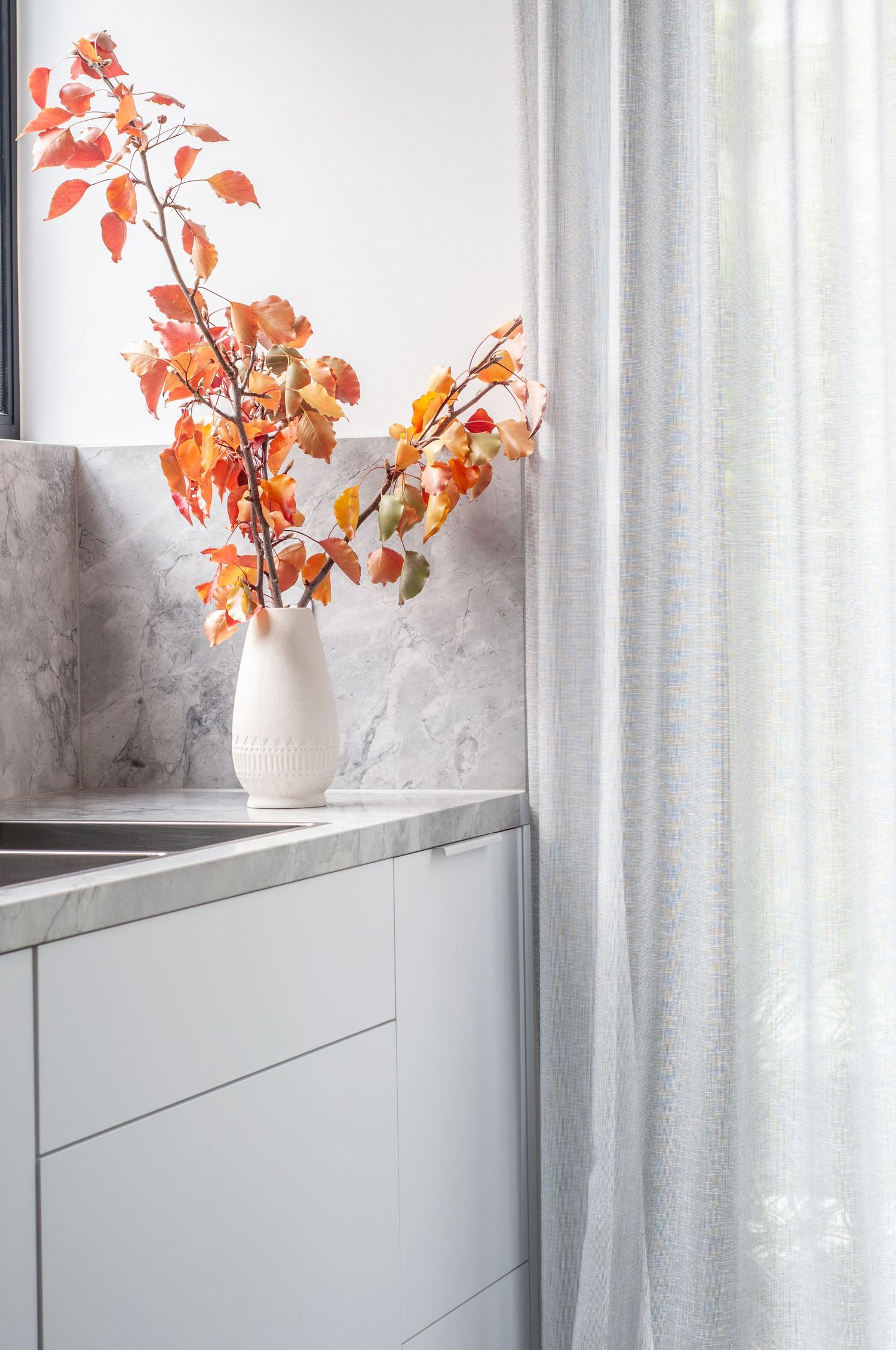 Orange autumn branches in white vase on marble countertop beside sheer curtains.
