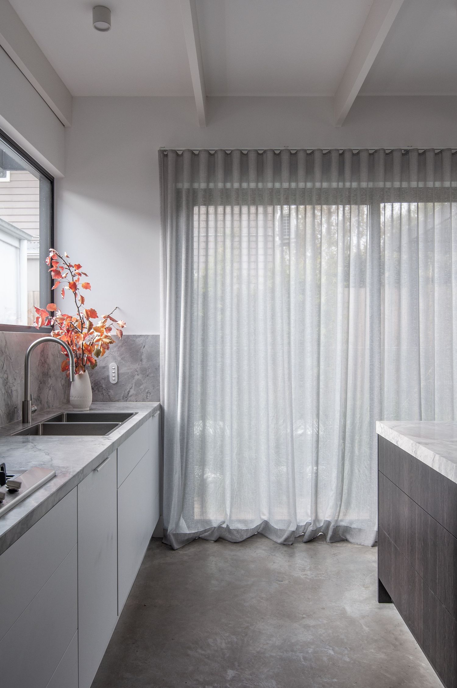 Modern white kitchen with gray marble counters and sheer curtains filtering natural light.