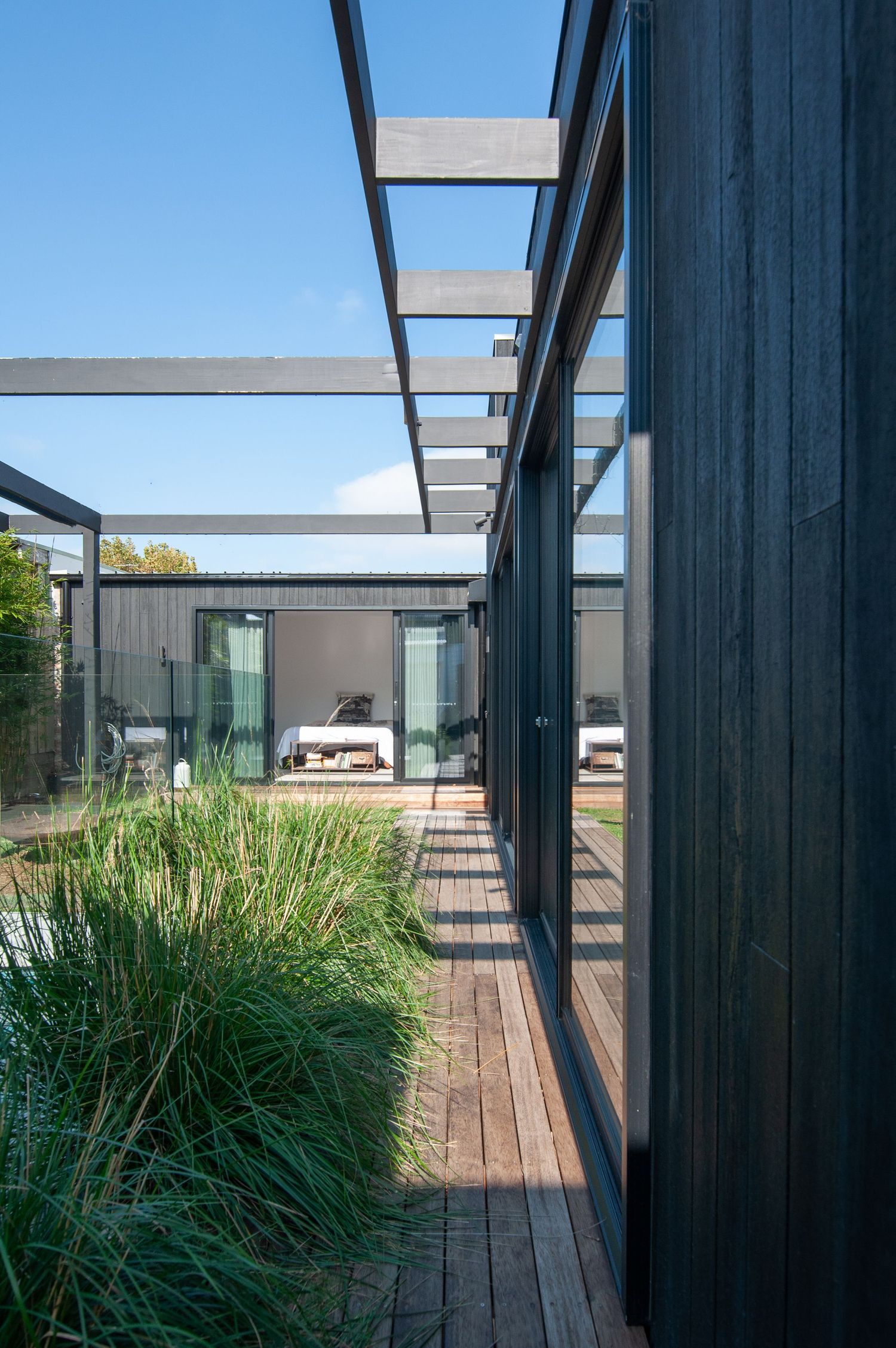 Covered walkway with wooden deck flooring and black structural beams leading to a modern house.
