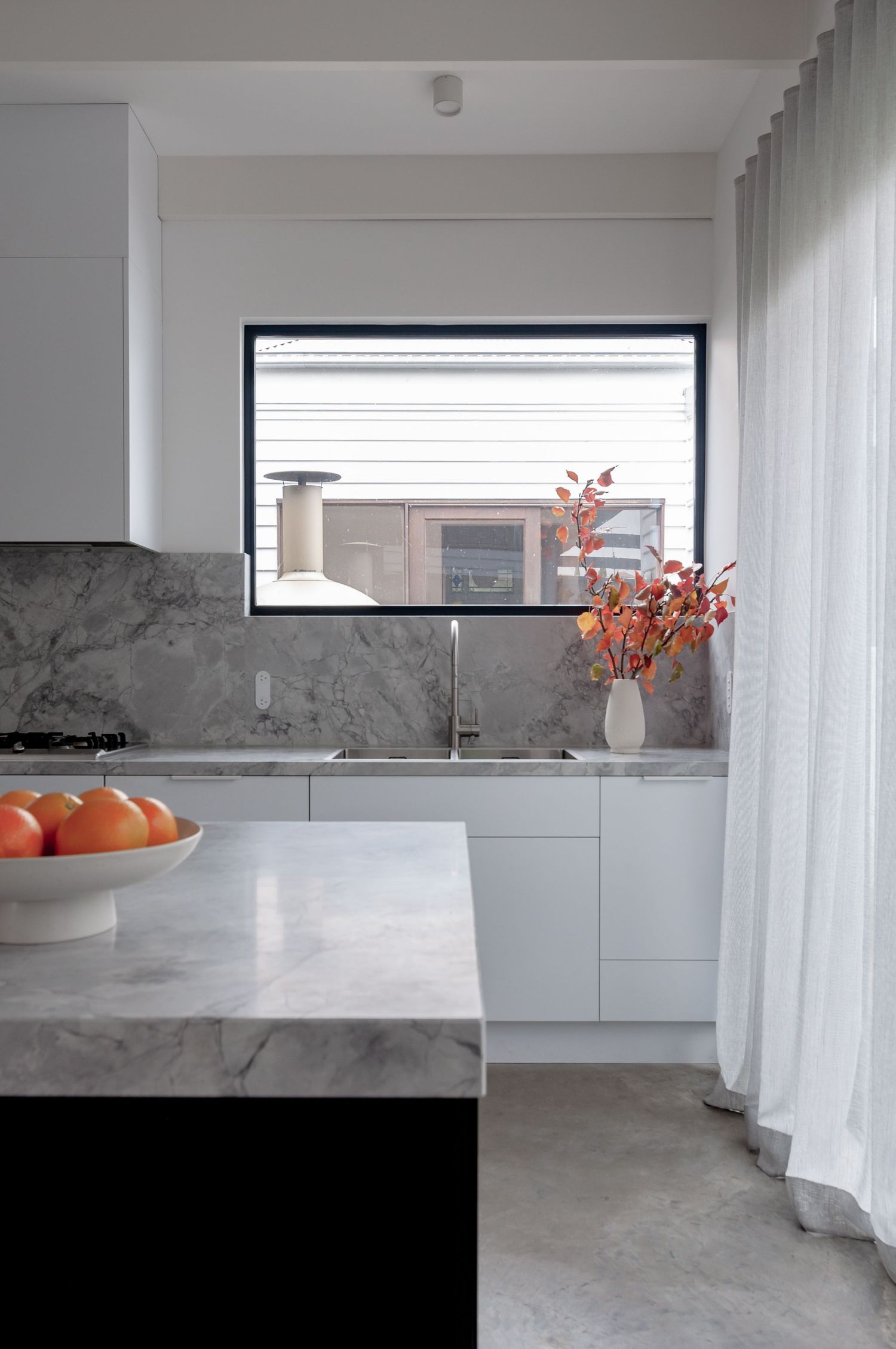 Modern white kitchen with marble countertops, fresh oranges, and orange floral arrangement by window.