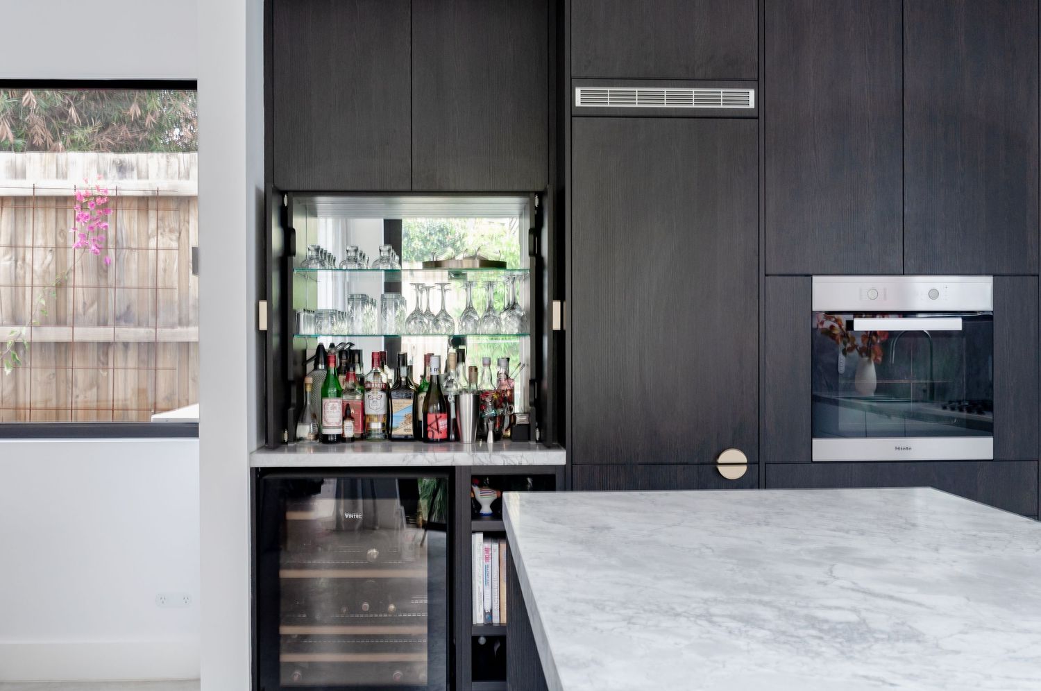 Hidden bar cabinet with glass shelving and liquor display in dark modern kitchen.