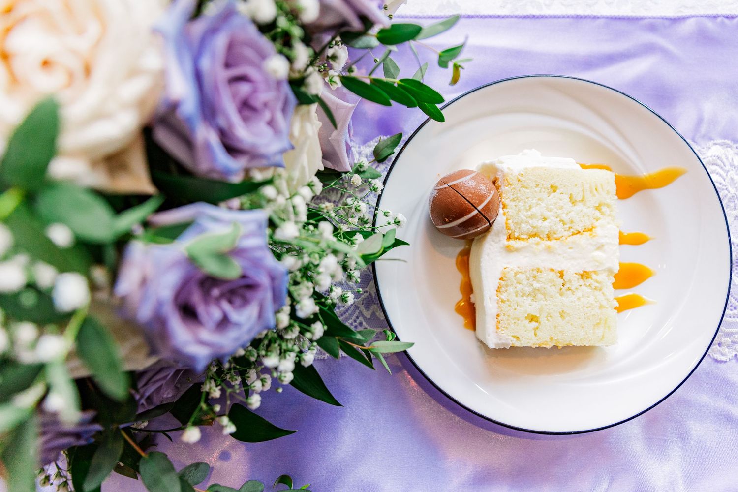 A slice of white wedding cake with purple roses and greenery decorating the table.