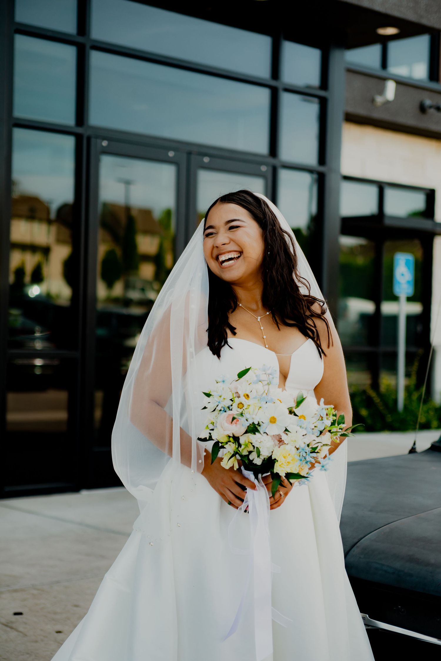 Radiant bride in flowing white dress holding white and yellow bouquet outside modern building.
