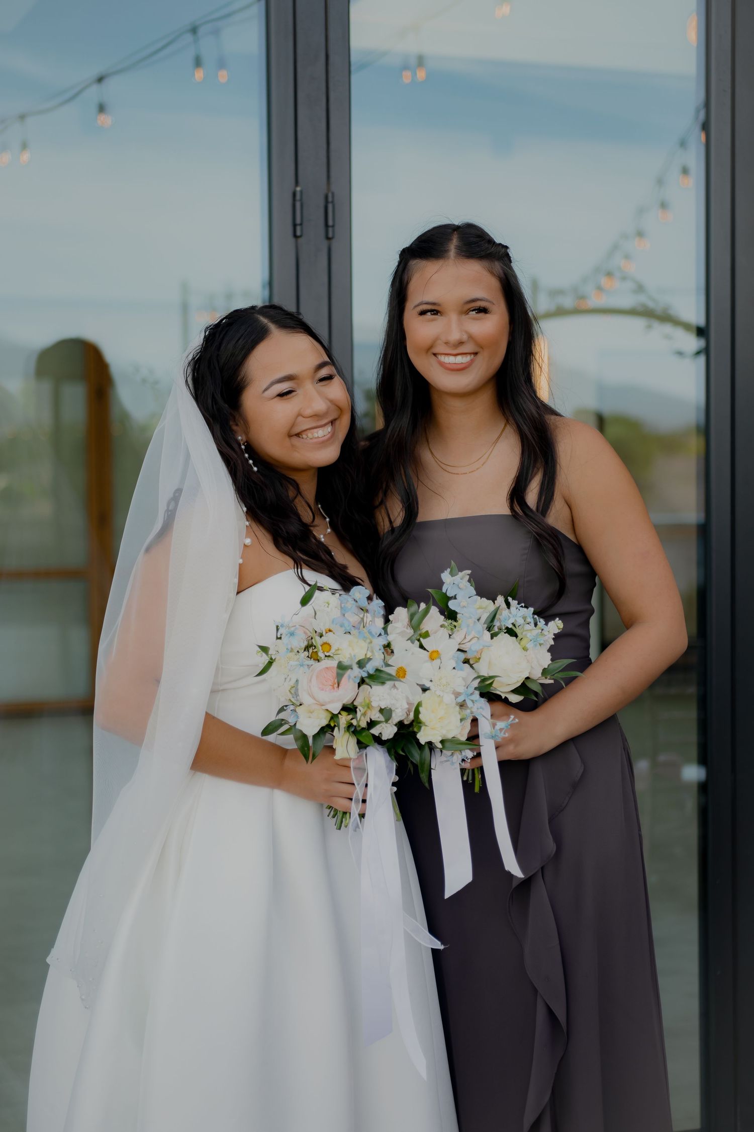 A bride and bridesmaid posing together with white floral bouquet against glass windows.