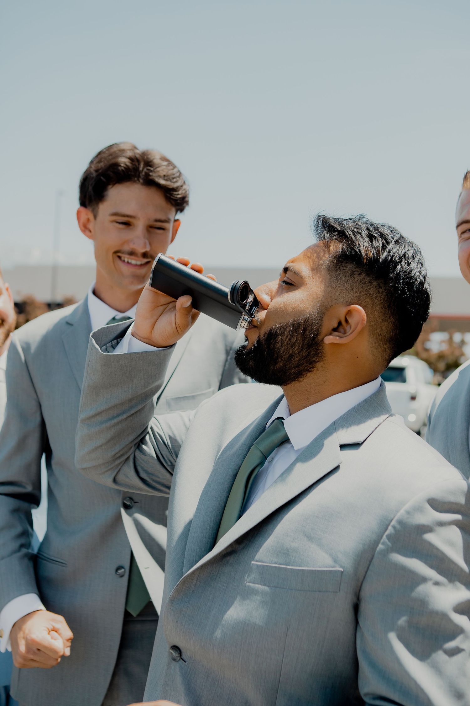 Groomsmen in light gray suits celebrating at outdoor wedding with drinks.