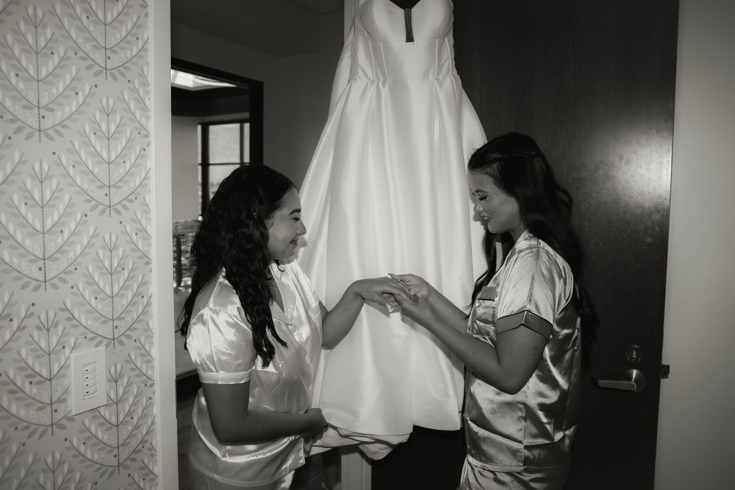 Two women examining white wedding dress details in preparation room.