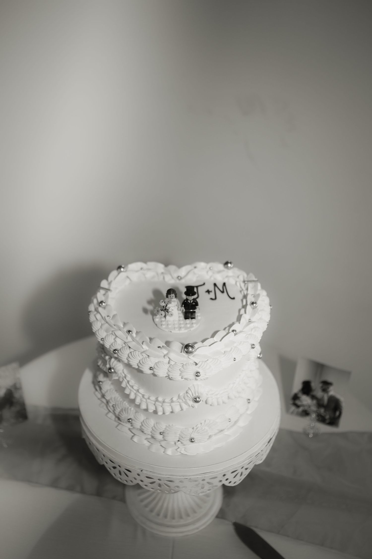 White two-tiered wedding cake with decorative piping and miniature bride and groom toppers on a pedestal stand.