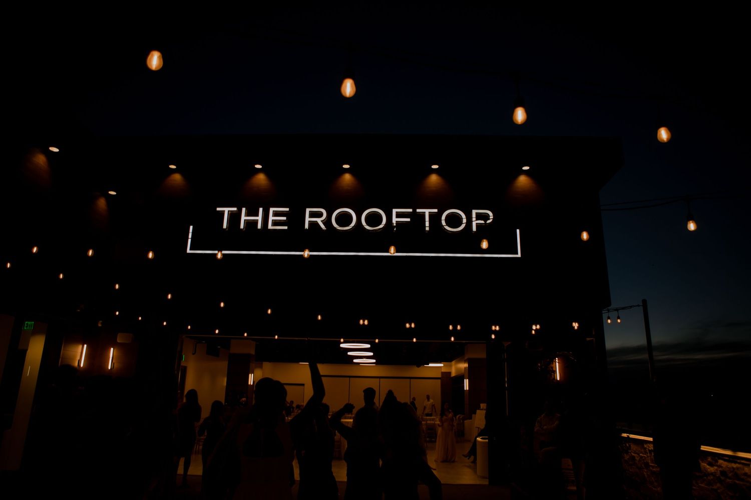 Illuminated rooftop venue entrance with string lights and silhouettes of people at dusk.
