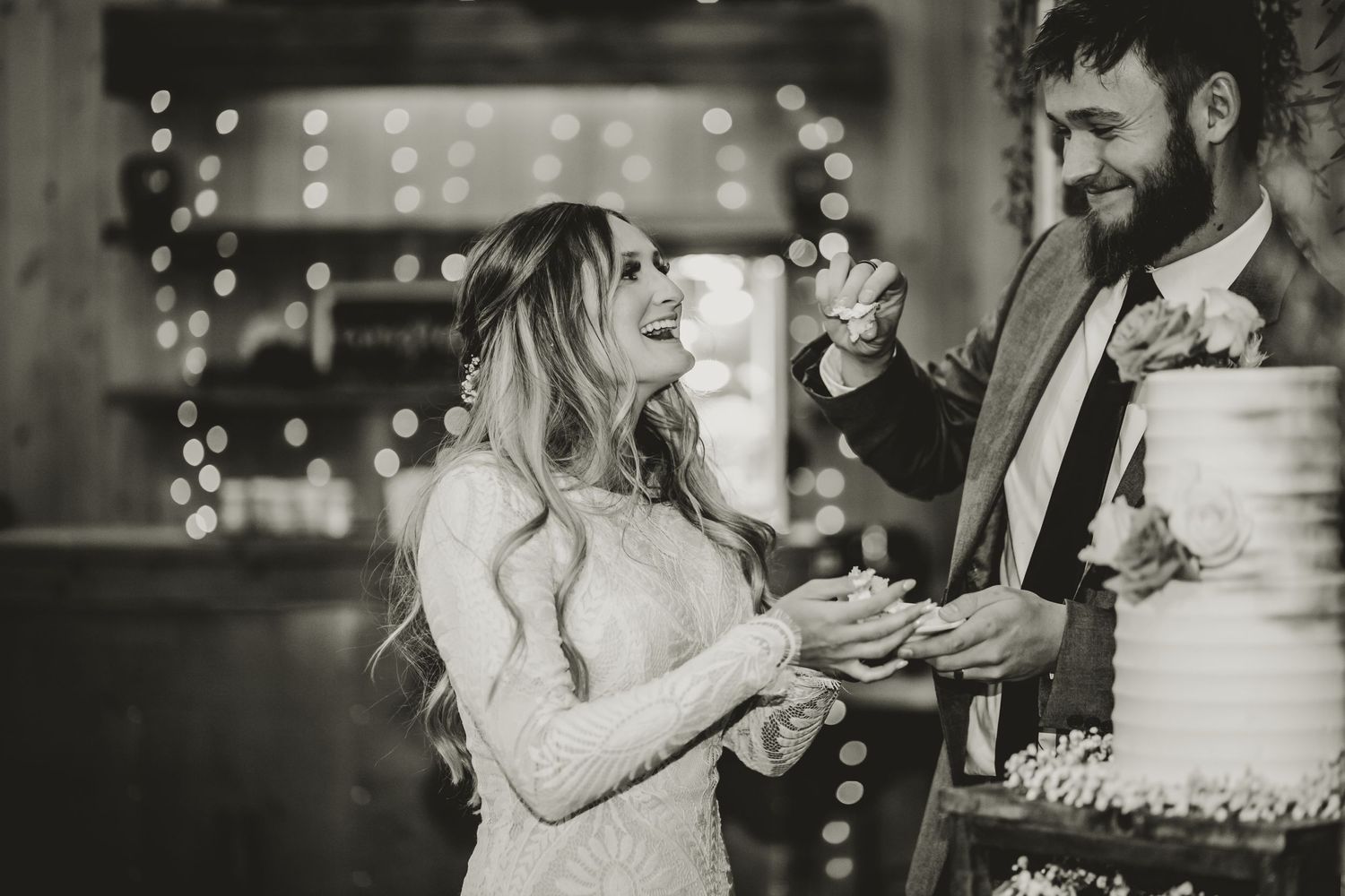 A black and white photo of a couple sharing cake at their wedding reception with twinkle lights in the background.