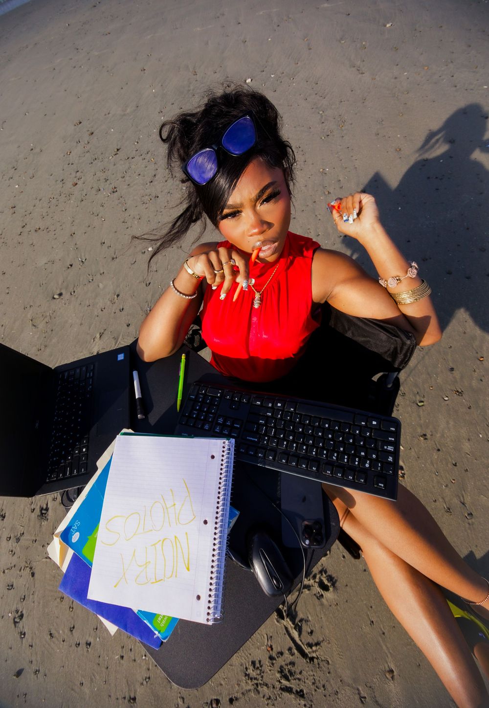 Woman in red top sits at outdoor desk on beach with keyboard, notebooks, and blue sunglasses.