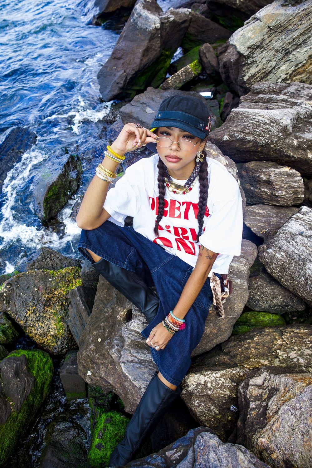 Young woman with braids in white crop tee and jeans sits on mossy rocks near ocean waves.