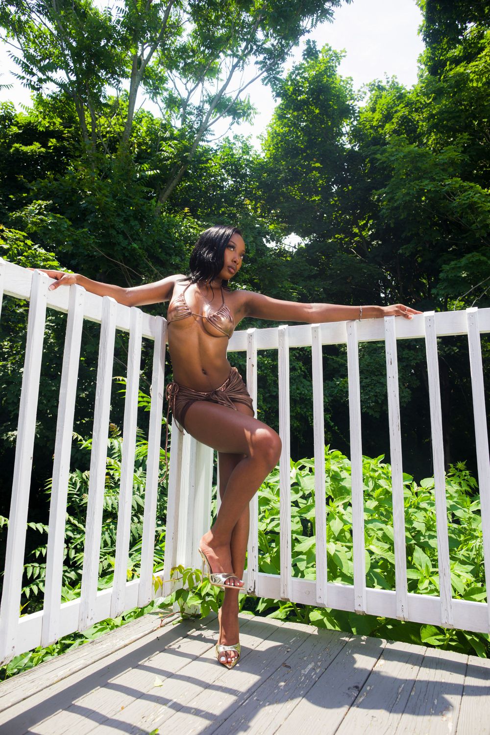 Woman in brown bikini posing on white deck railing surrounded by green trees.