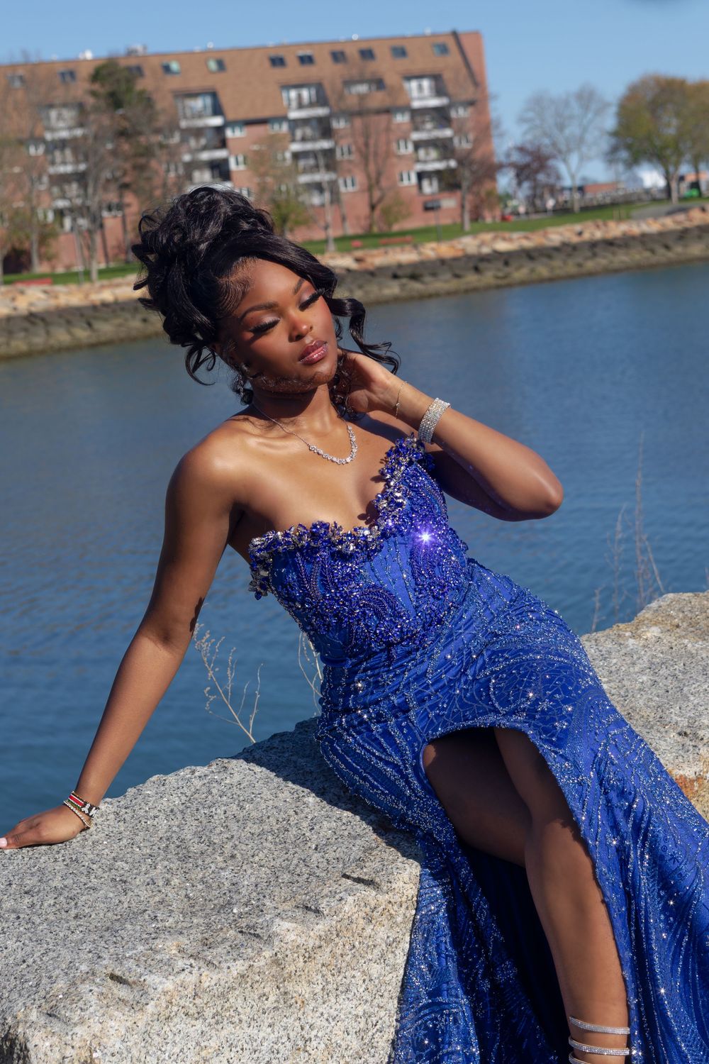 Young woman in a royal blue sequined mermaid gown posing on rocks by a waterfront on a sunny day.