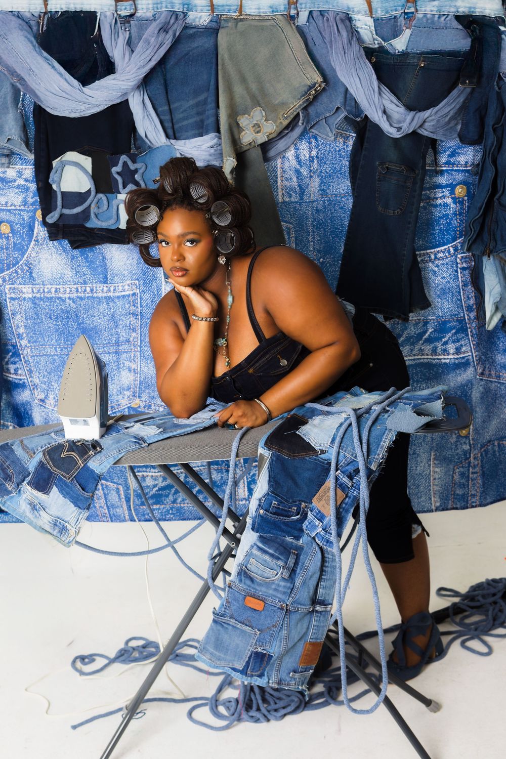Woman with hair rollers posing at an ironing board surrounded by denim clothing in a creative studio setup.