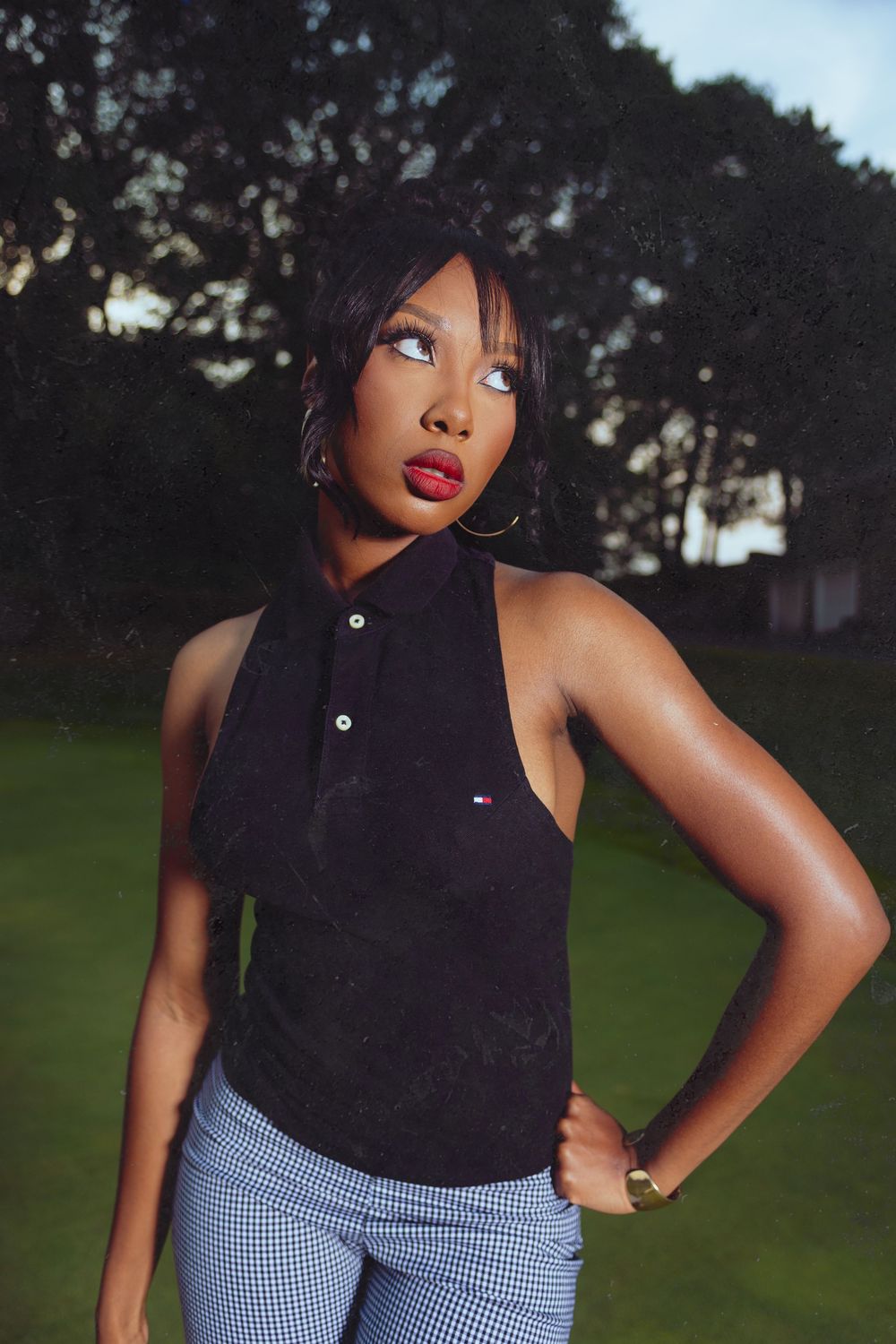 Stylish woman in a black sleeveless Tommy Hilfiger polo and plaid pants posing outdoors at dusk.