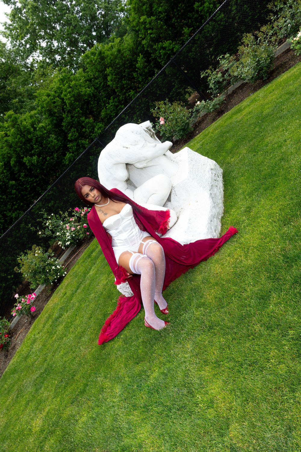 Woman in white lingerie and red cape reclines on grass near white sculpture in garden.