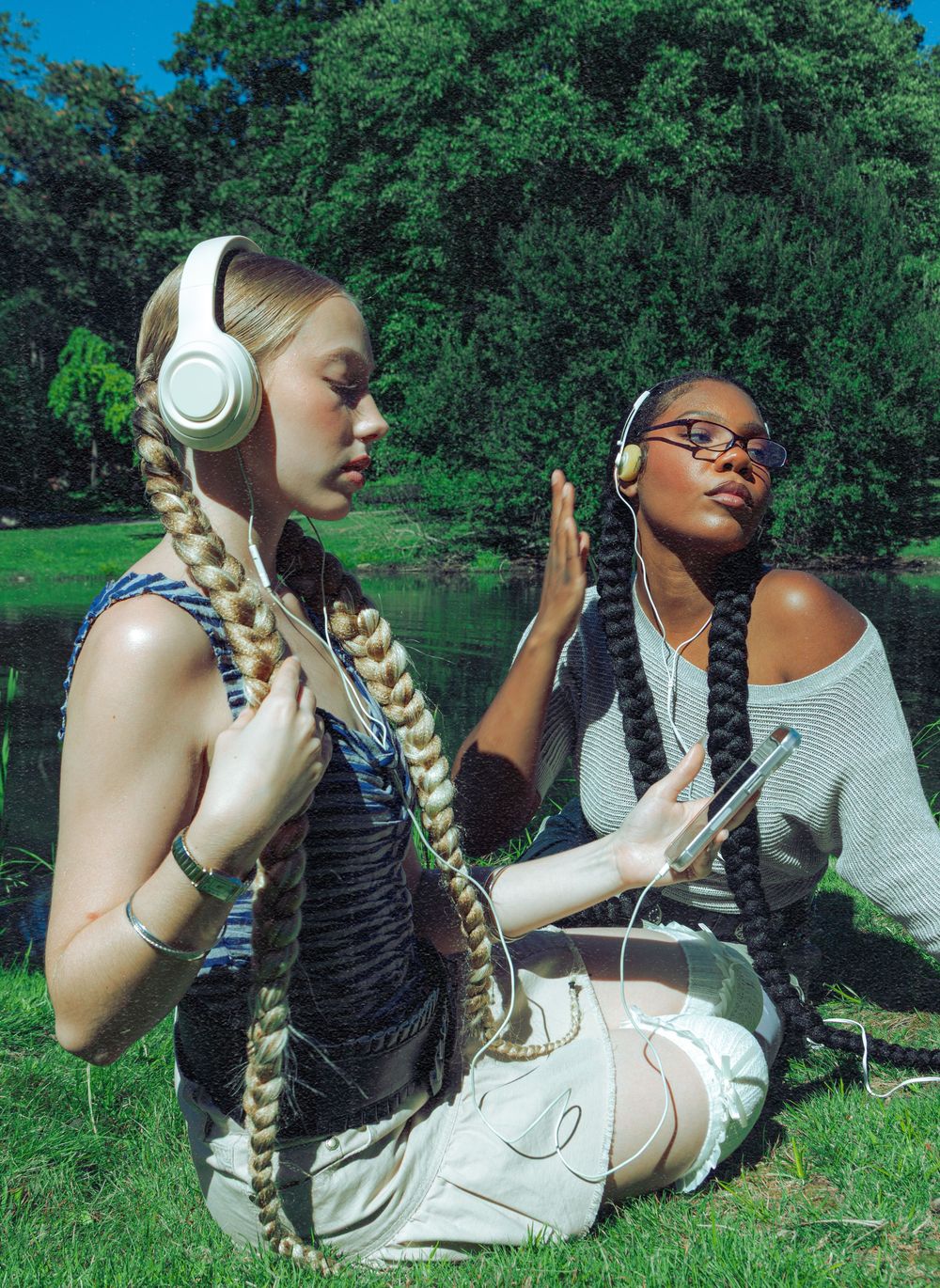 Two women with long braids wearing headphones sit on grass in park by a pond.